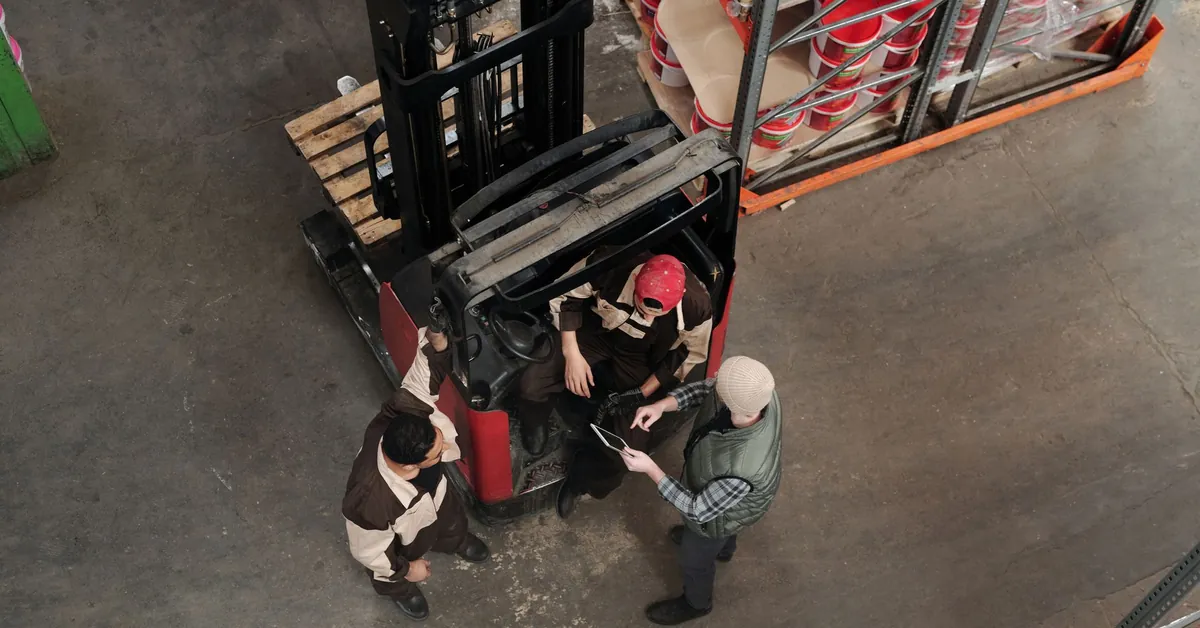 Top view of workers discussing plans in a warehouse beside a forklift, emphasizing teamwork. employees talking with each other