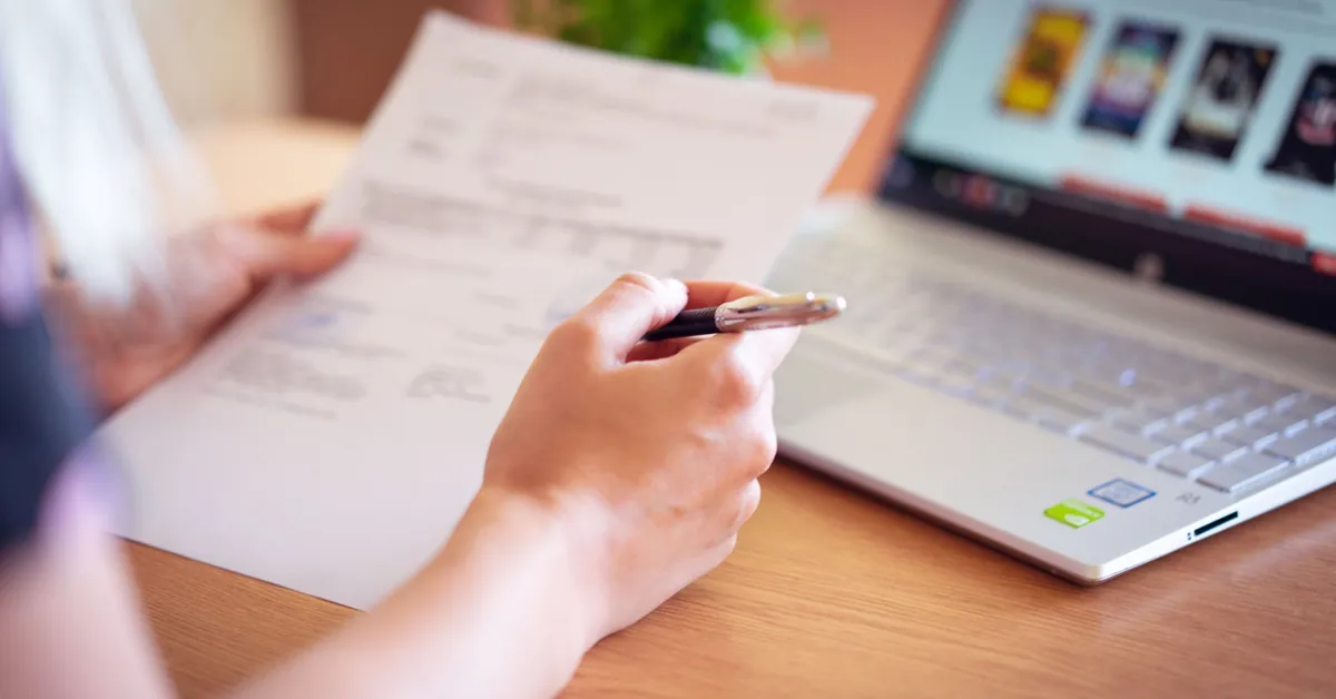 Close-up of a hand holding a pen and paper near a laptop, suggesting online shopping or document review. person holding pen and paper