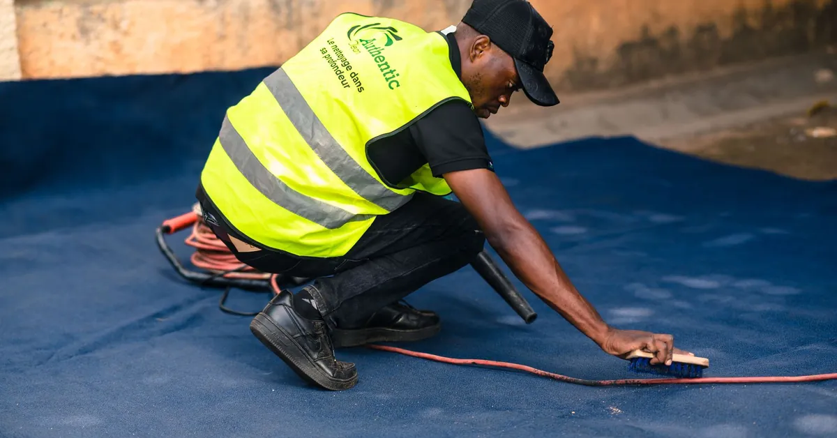 A worker dressed in a safety vest cleans a blue carpet outdoors with a brush. worker cleaning carpet with brush in outdoor setting
