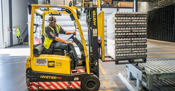 A warehouse worker maneuvers a forklift to transport crates for brewing company storage. person using forklift