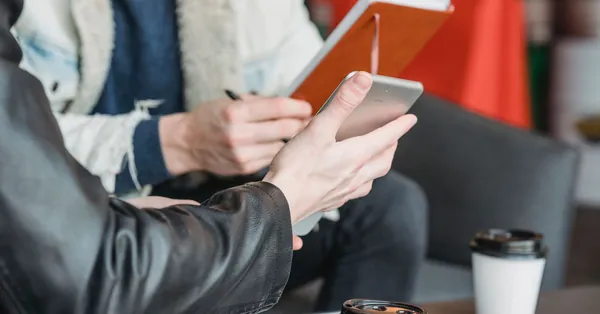 Crop anonymous man showing tablet to male partner sitting at table with notebook and coffee cups during work in cafe colleagues sharing tablet with working in coffee shop