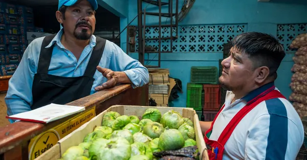 Two men in a market discussing vegetables. Fresh produce in a box nearby. two market vendors with fresh produce indoors