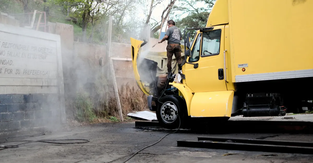 A mechanic inspects and repairs the engine of a yellow truck in an outdoor setting, surrounded by steam. a yellow truck with a man spraying water on it