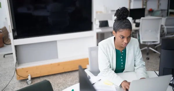 Focused woman using a laptop in a modern office setting, wearing a white blazer, working diligently. woman in white blazer sitting at table using laptop