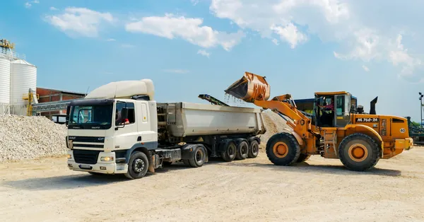 Excavator loading materials into a heavy-duty truck at a sunny construction site. brown loader beside white cargo truck