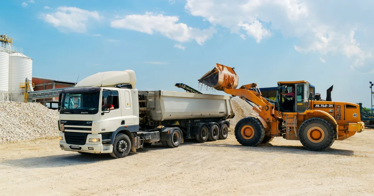 Excavator loading materials into a heavy-duty truck at a sunny construction site. brown loader beside white cargo truck